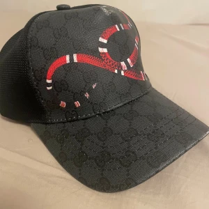 Svart keps med röd detalj från Gucci - Snygg svart keps från Gucci med ett coolt rött ormmönster på framsidan. Kepsen har ett justerbart band baktill med blå och röda ränder. Perfekt för att ge din outfit en lyxig touch.
