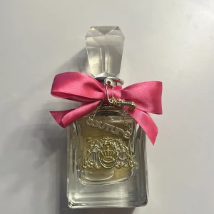 Parfym med rosa rosett och gulddetaljer - Säljer en lyxig parfym från Juicy Couture. Flaskan är elegant med en stor, facetterad glaskork och dekorerad med en iögonfallande rosa rosett. Den har också guldfärgade detaljer och en charm med texten 'couture'. Perfekt för den som älskar en glamorös och feminin stil.