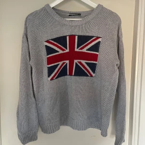 Grå stickad tröja med Union Jack-motiv - Säljer en snygg grå stickad tröja med en Storbritannien flagga på framsidan. Tröjan är perfekt för höst och vinter med sina långa ärmar och ribbade kanter. Den är i bra skick och supermysig att ha på sig. Passar perfekt för en avslappnad stil!