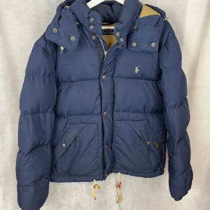 Blå dunjacka från Ralph Lauren - Säljer en snygg blå dunjacka från Ralph Lauren. Jackan har en fet design med dragkedja och knappar framtill. Den är långärmad och perfekt för vintern. Även  justerbar luva. Priset är inte hugget i sten ! 