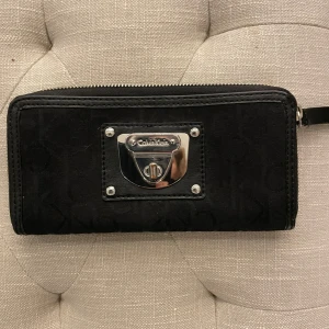 Svart plånbok från Calvin Klein - Säljer en snygg svart plånbok från Calvin Klein. Den har en stilren design med en silverfärgad metallplatta på framsidan där märket är ingraverat. Plånboken har en dragkedja och flera fack inuti för kort och kontanter. Nyskick