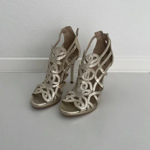 Guldiga klackskor från Cobler - Säljer ett par snygga guldiga klackskor från Cobler. De har en öppen tå och ett coolt flätat mönster som ger en elegant look. Perfekta för fest eller bal! Skorna har en hög klack och är i nyskick.