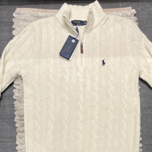 Vit kabelstickad tröja från Ralph Lauren - Säljer en stilren vit kabelstickad tröja från Ralph Lauren med dragkedja och broderad logga på bröstet. Perfekt för kyliga dagar med sin höga krage och mjuka material. Storlek: L men passar M