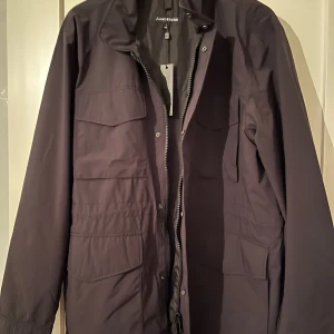  J. Lindeberg jacka - Snygg svart jacka från J. Lindeberg med dragkedja och knappar. Jackan har långa ärmar och flera praktiska fickor framtill. Perfekt för en stilren look.