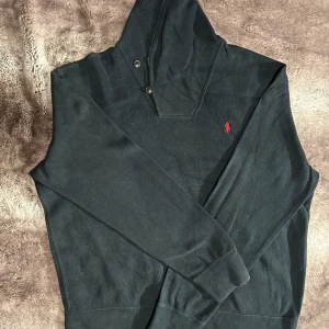 Polo Ralph Lauren - Säljer en stilren svart hoodie från Polo Ralph Lauren med det klassiska röda logotypen på bröstet. Tröjan har långa ärmar och en praktisk huva med knappar. Perfekt för en avslappnad stil.