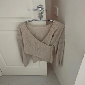 Beige ribbad omlottopp - Säljer en stilren beige omlottopp med ribbad struktur. Toppen har långa ärmar och en v-ringad design som ger en elegant look. Perfekt för både vardag och speciella tillfällen.