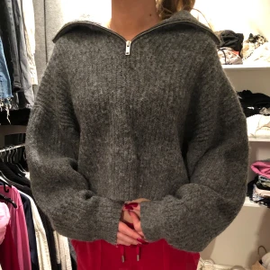 Grå stickad tröja med dragkedja - Mysig grå stickad tröja med dragkedja och hög krage. Perfekt för kyliga dagar med sin croppade design och långa ärmar. En stilren och bekväm tröja för alla tillfällen.