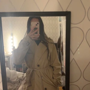Beige trenchcoat - Snygg beige trenchcoat med knytskärp i midjan och klassisk krage. Perfekt för höst och vår. Långärmad design med knappar framtill.