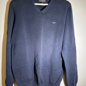 Mörkblå tröja från Hampton Republic - Säljer en stilren mörkblå tröja från Hampton Republic med v-ringning och ett litet broderat emblem på bröstet. Tröjan har långa ärmar och ribbade kanter för en klassisk look. Perfekt för en avslappnad men ändå snygg stil.