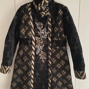 Mönstrad kappa från Desigual - Unik kappa från Desigual med ett iögonfallande mönster i svart och beige. Kappan har långa ärmar och knappar framtill. Den är dekorerad med broderade blommor och har en hög krage för extra stil.stl 42 men liten i stoleken, som en stl 40. Nyskick använd 1 ggr några timmar. 