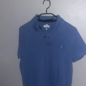 Blå pikétröja från Hollister - Säljer en blå pikétröja från Hollister med deras ikoniska logga på bröstet. Tröjan har en klassisk krage och knappar framtill. Perfekt för en avslappnad stil.