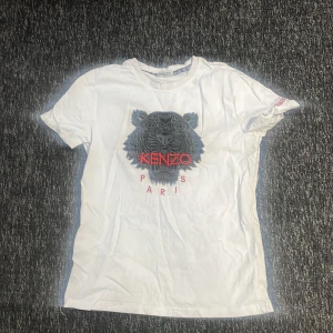 Vit t-shirt från Kenzo med tigertryck - Snygg vit t-shirt från Kenzo med ett coolt tigertryck i grått och rött på framsidan. T-shirten har korta ärmar och är perfekt för en avslappnad stil. Kenzo-loggan är tydligt synlig i rött.