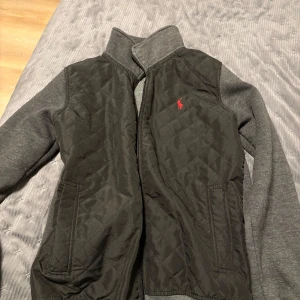 Grå jacka från Polo Ralph Lauren - Snygg grå jacka från Polo Ralph Lauren med quiltad framsida och broderad logga i rött. Jackan har långa ärmar och dragkedja framtill. Perfekt för kyliga dagar.