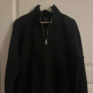 Svart tröja med dragkedja från H&M - Säljer en svart tröja från H&M i relaxed fit med dragkedja framtill. Perfekt för en avslappnad stil. Tröjan har långa ärmar och en hög krage.