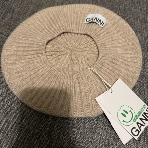 Beige ull mössa från Ganni - Snygg beige stickad basker från Ganni med ribbad design. Perfekt för att hålla värmen under kyliga dagar. Märkt med Ganni's 'Responsible' etikett för hållbarhet.