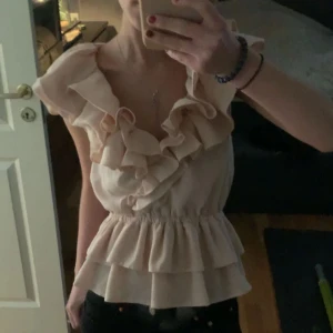 Ljus rosa blus med volangdetaljer - Säljer en söt ljus rosa blus med volangdetaljer. Blusen har korta ärmar och en v-ringad design. Perfekt för en stilren och feminin look.