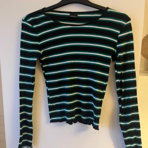 Randig långärmad tröja från Gina Tricot - Säljer en randig långärmad tröja från Gina Tricot i svart, vit och grön. Tröjan har en rund halsringning och är ribbad för en bekväm passform. Perfekt för en avslappnad stil.