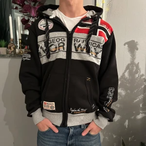 Svart och grå hoodie från Geographical Norway - Snygg svart och grå hoodie med dragkedja från Geographical Norway. Den har tryck och broderade detaljer i vitt och rött samt en praktisk huva med snörning. Perfekt för en avslappnad stil.