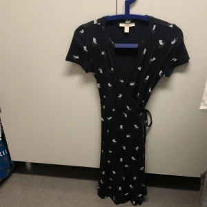 Mörkblå omlottklänning från Esprit - Säljer en mörkblå omlottklänning från Esprit med korta ärmar och ett mönster av små solsstolar. Klänningen har en knytning i midjan och slutar över knäna.