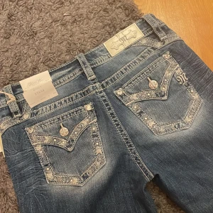 Blå jeans från Miss Me - Jag är 173 och bär oftast 38/40. Pris kan diskuteras. Helt nya , aldirg använda. Snygga blå jeans från Miss Me med dekorativa bakfickor och broderade detaljer. Byxorna har en bootcut-stil och är midwaist. Perfekta för en avslappnad look med en touch av stil.