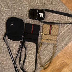 Fyra axelväskor i olika stilar - Säljer fyra axelväskor i olika stilar och färger. En svart väska med dragkedja och justerbar rem, en glansig svart med vit logga, en svart med röd-grön rand och en beige med monogrammönster och gulddetaljer. Perfekta för olika tillfällen och outfits. Köp alla för 150kr 