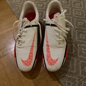 Vita Nike fotbollsskor med röda detaljer - Säljer ett par vita Nike fotbollsskor med röda och svarta detaljer. Skorna har en stor Nike-logga på sidan och snörning framtill. Sulan är röd med gula dobbar, perfekt för bra grepp på planen.