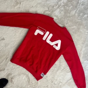 Röd sweatshirt från FILA - Säljer en snygg röd sweatshirt från FILA med stort vitt logotryck på framsidan. Tröjan är tillverkad i en bomulls- och polyesterblandning och har långa ärmar. Perfekt för en sportig look!