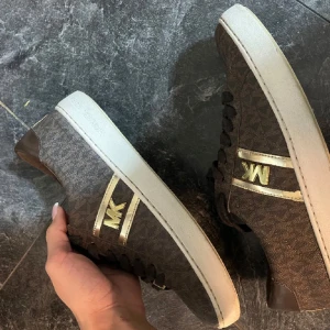 Bruna sneakers från Michael Kors - Snygga bruna sneakers från Michael Kors med gyllene detaljer och logotyp. Skorna har en vit sula och snörning framtill. Perfekta för en stilren look. Stl 39, köpta på nk. Använt få gånger och nu inte kommer till användning. Nypris 2200kr