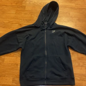Mörkblå hoodie från Gymshark - Säljer en mörkblå hoodie från Gymshark med dragkedja och huva. Tröjan har en liten vit logotyp på bröstet och praktiska fickor framtill. Perfekt för en avslappnad stil eller träning.