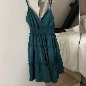 Grön ärmlös klänning - Snygg mörk grön/blå klänning med smala axelband och omlott-design. Den har en smockad midja och volanger som ger en fin siluett. Perfekt till sommaren!!🤩🤩🩷🩷