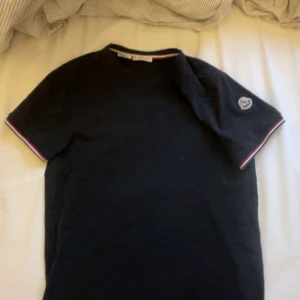 Svart t-shirt från Moncler - Snygg svart t-shirt från Moncler med korta ärmar och en broderad logga på ärmen. Den har en stilren design med ränder i rött och vitt vid ärmslut och halsringning.