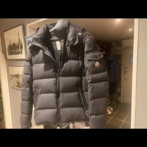 Mörkgrå dunjacka från Moncler - Snygg mörkgrå dunjacka från Moncler med hög krage och dragkedja. Jackan har långa ärmar och en broderad logga på ärmen. Perfekt för kyliga dagar. Det negativa är att den tecknade bilden på insidan behöver sys då den sitter halvt löst därav priset