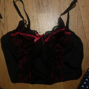 Svart och röd korsett med spetsdetaljer - Elegant svart korsett med röda detaljer och spets. Justerbara axelband och en söt rosett framtill. Perfekt för en speciell kväll.