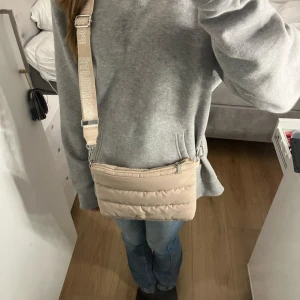 Beige axelväska från H&M Divided - Snygg beige axelväska från H&M Divided med quiltad design och justerbar axelrem med texten 'Possibilité'. Perfekt för att bära dina nödvändigheter med stil.