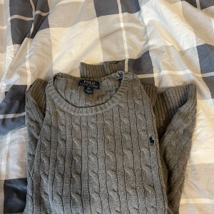 Grå stickad tröja från Polo Ralph Lauren - Säljer en stilren grå stickad tröja från Polo Ralph Lauren med kabelstickat mönster. Tröjan har långa ärmar och en liten broderad logotyp på bröstet. Perfekt för kyligare dagar. Är i storlek xl men sitter som m
