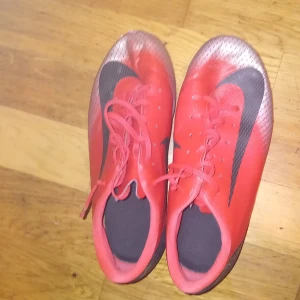 Röda fotbollsskor från Nike - Säljer ett par röda Nike fotbollsskor med svarta detaljer och snörning. Skorna har dobbar för bra grepp på planen och en stilren design med Nike-loggan på sidan.
