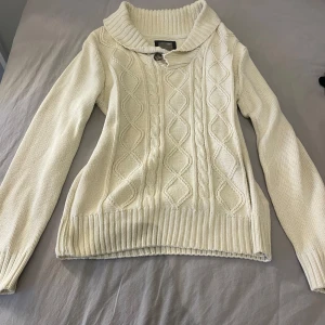 Beige stickad tröja från Cubus - Säljer en mysig beige stickad tröja från Cubus med ett snyggt kabelstickat mönster. Tröjan har en hög krage med knappdetalj och långa ärmar. Perfekt för kyliga dagar!