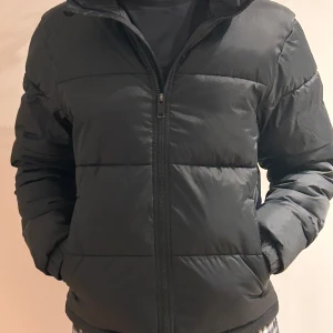Svart pufferjacka från Everest - Säljer en svart pufferjacka från Everest i storlek XXS. Jackan har en dragkedja framtill och är perfekt för kyligare väder. Har ingen luva. Den är quiltad och har långa ärmar för extra värme.