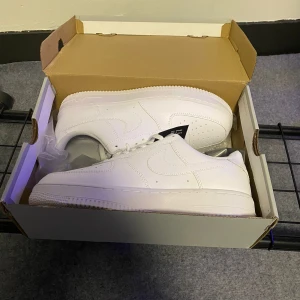 Vita Nike Air Force 1 - Säljer ett par klassiska vita Nike Air Force 1 sneakers. De har en stilren design med snörning och är tillverkade i syntetmaterial. Perfekta för en sportig och trendig look.