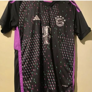 Svart och lila FC Bayern München fotbollströja - Snygg svart fotbollströja från Adidas med lila och gröna detaljer. FC Bayern München-logga på bröstet och text på ryggen. Perfekt för fans som vill sticka ut på läktaren eller planen.