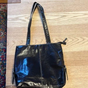 Svart axelväska från LBVYR - Snygg svart axelväska i syntetmaterial från LBVYR. Väskan har dubbla handtag och en dragkedja upptill för säker förvaring. Perfekt för att bära dina dagliga nödvändigheter med stil.