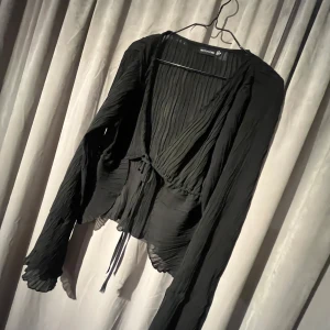 Svart blus från PrettyLittleThing, 36/S - Snygg svart ribbad blus från PrettyLittleThing med långa ärmar och knytning framtill. Perfekt för en stilren look med en touch av elegans. Blusen har en luftig och bekväm passform.