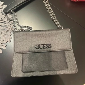 Svart axelväska från Guess - Snygg svart axelväska från Guess med kedjeaxelrem och logotyp framtill. Väskan har en praktisk dragkedja inuti och en magnetknapp för enkel stängning. Perfekt för att bära dina nödvändigheter med stil. Säljes då den inte kommer till användning. Nypris ca 1800kr säljes för 1000kr 