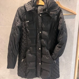 Svart quiltad jacka från Rockandblue - Snygg svart quiltad jacka från Rockandblue med dragkedja och knappar. Jackan har flera praktiska fickor med dragkedjor och en stilren design. Perfekt för kyligare dagar. Säljer nu denna på grund av att den är lite för liten i storlek. Vet tyvärr inte exakta storleken men gissar på S/M. 