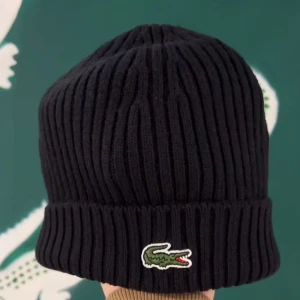 Svart ribbad mössa från Lacoste - Snygg svart ribbad mössa från Lacoste med det ikoniska krokodilemblem broderat framtill. Perfekt för att hålla värmen under kyliga dagar.