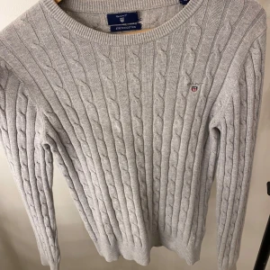 Grå stickad tröja från GANT - Säljer en stilren grå stickad tröja från GANT i stretchig bomull. Tröjan har ett klassiskt kabelstickat mönster och rund halsringning. Perfekt för kyligare dagar. Den passar som storlek S också 