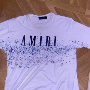 Vit t-shirt från Amiri med stänkmönster - Säljer en vit t-shirt från Amiri med ett coolt stänkmönster i blått och svart. T-shirten har korta ärmar och en avslappnad passform. Perfekt för en trendig look. Pris kan diskuteras vid snabb affär!