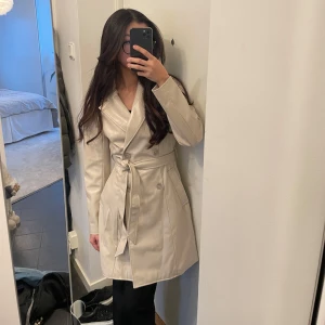 Beige skinn trenchcoat från H&M Divided - Snygg beige skinn trenchcoat från H&M Divided med knytbälte i midjan och knappar framtill. Strl Xs och har använt den få ggr.