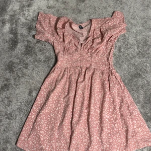 Rosa blommig klänning - Charmig rosa klänning med blommigt mönster och korta ärmar. Den har en smickrande passform med rynkade detaljer vid bysten och en v-ringad hals. Perfekt för en somrig look!
