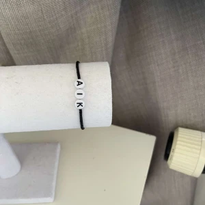 Svart armband med bokstäver - Säljer ett enkelt och stilrent svart armband med vita pärlor som har bokstäverna 'A', 'I', 'K'. Perfekt för att visa ditt stöd eller bara som en snygg accessoar.